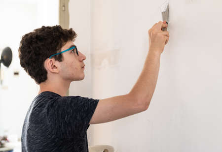 Masonry work in the apartment: a Caucasian boy is intent on filling the imperfections of the wall. On the scraper there is a dose of putty paste that is spread on the wall.の写真素材