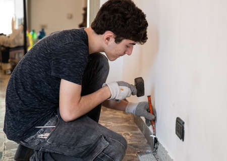 Masonry work in the apartment: a Caucasian boy is wearing work gloves and is using hammer and chisel to demolish the old baseboard.の写真素材