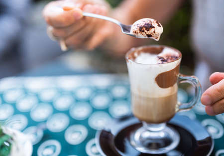 An inviting mocha: close-up shot on the spoon that raised the creamy foam with cocoa powder on it. In the glass the colors of milk, coffee, chocolate cream form a pleasant pattern.の写真素材