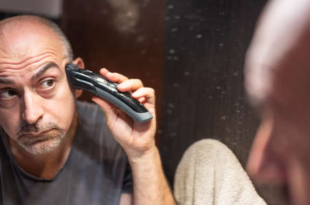 A middle-aged Caucasian man cuts his hair in the bathroom. While placing the clipper on your head check with your eyes in the mirror.の写真素材