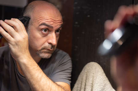 A middle-aged Caucasian man cuts his hair in the bathroom. While placing the clipper on your head check with your eyes in the mirror.の写真素材