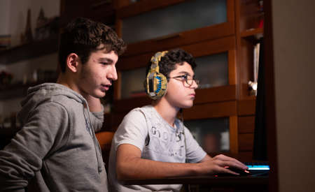 Staying at home: Two Caucasian brothers keep busy with their favorite video game. One is sitting with the gaming headset on his head and the other is standing next to him to suggest moves to take.の写真素材