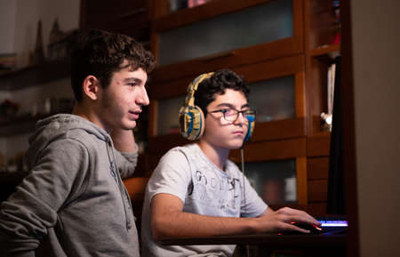 Staying at home: Two Caucasian brothers keep busy with their favorite video game. One is sitting with the gaming headset on his head and the other is standing next to him to suggest moves to take.の写真素材