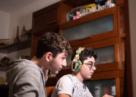 Staying at home: Two Caucasian brothers keep busy with their favorite video game. One is sitting with the gaming headset on his head and the other is standing next to him to suggest moves to take.の写真素材