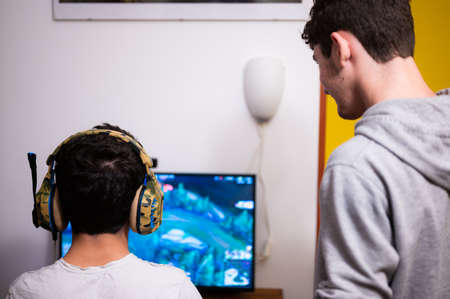 Staying at home: Two Caucasian brothers keep busy with their favorite video game. One is sitting with the gaming headset on his head and the other is standing next to him to suggest moves to take.の写真素材