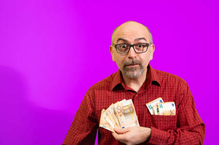Middle-aged Caucasian man holding a bunch of 50 euro bills in his hand and has more in his shirt pocket. He is happily surprised. Concept of winning, luck and wealth. Yellow background.の写真素材