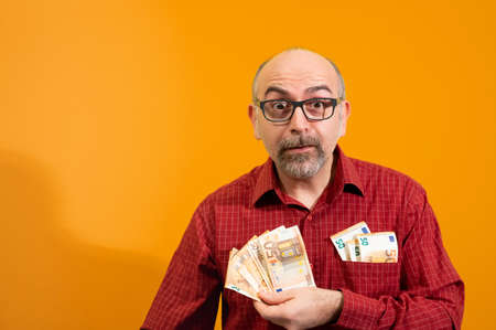 Middle-aged Caucasian man holding a bunch of 50 euro bills in his hand and has more in his shirt pocket. He is happily surprised. Concept of winning, luck and wealth. Yellow background.の写真素材