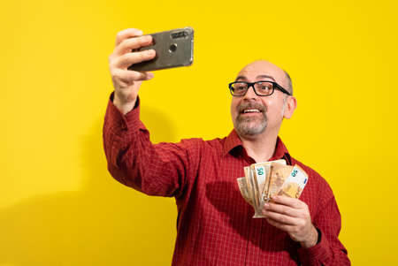 Middle-aged Caucasian man holding a bunch of 50 euro bills in one hand and taking a selfie with the other. He is happily surprised. Concept of winning, luck and wealth. Yellow background.の写真素材