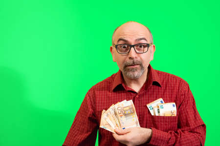 Middle-aged Caucasian man holding a bunch of 50 euro bills in his hand and has more in his shirt pocket. He is happily surprised. Concept of winning, luck and wealth. Yellow background.の写真素材