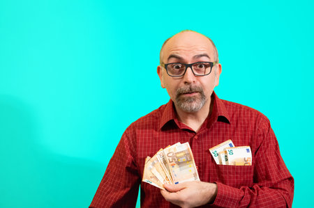 Middle-aged Caucasian man holding a bunch of 50 euro bills in his hand and has more in his shirt pocket. He is happily surprised. Concept of winning, luck and wealth. Yellow background.の写真素材