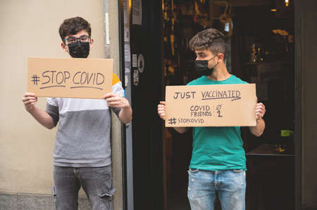 Two friends with masks display two signs with the message stop covid, just vaccinated. To underline...