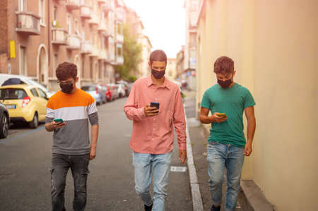 Friends group walking on city street with protective black face mask using the smartphone. Pandemic time. New normal lifestyle concept with young people covered by protective mask. Blurred background.の写真素材