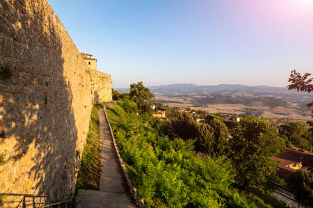 The walls of the historic village. The warm light of the late afternoon enhances the massive stone wall, at its foot the perimeter path. Tuscan landscape.の写真素材