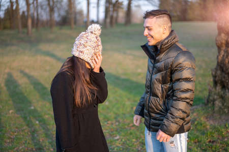 Caucasian couple in casual wear is in the spring park. Happy and smiling they joke among themselves: he pretends to puddle her and with a quick movement of her he lowers her cap covering her face.の写真素材