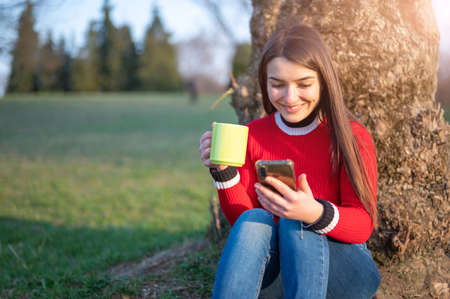 An attractive young brunette woman is sitting at the foot of a tree at the park. She gets a message, drinks from the green cup. Concept of carefree, communication use technologyの写真素材