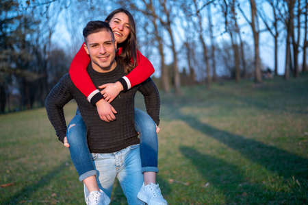 Caucasian couple in spring park smiling, attractive and happy girl hugs handsome athletic guy from shoulders. He moves hloding her piggyback, couple outdoors.の写真素材