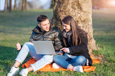 A beautiful couple in casual wear is having fun with the laptop outdoors. Sitting at the foot of a...