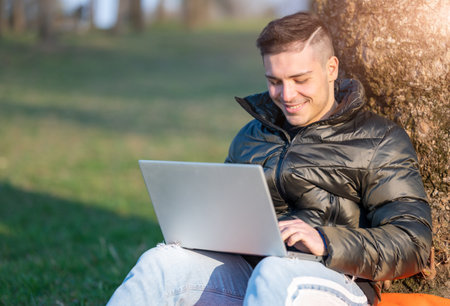 Focused handsome man in casual wear working with laptop outdoors. Sitting at the foot of a tree, he enjoys nature on a sunny spring day. Remote working concept. Copy space.の写真素材
