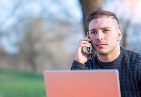 Portrait of attractive young office worker in casual wear, he received a phone call on his cellphone, he is concentrated while listening. Concept of working remotely with laptop outdoors in a park.の写真素材