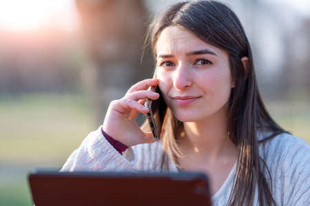 Portrait of a young beautiful woman working remotely in a park, she is focused while talking on her cell phone. In front of her, her tablet. Remote working outdoors concept.の写真素材