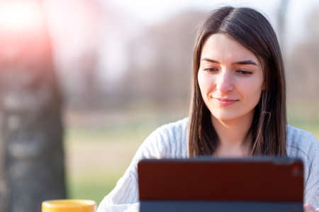 Portrait of a smiling young beautiful woman working remotely in a park. In front of her, her tablet and her yellow cup of coffee. Beautiful spring day. Remote work outdoors concept.の写真素材