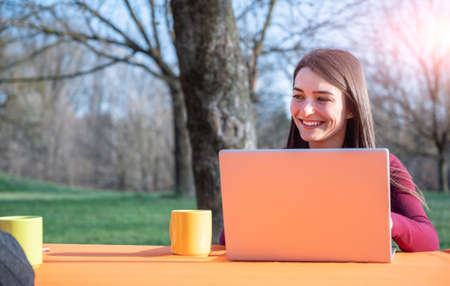 Portrait of young woman works remotely in a park on a nice sunny day. She use a laptop  while talking to each other. On the table are cups. Remote working concept outdoors.の写真素材