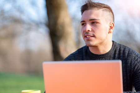Portrait of attractive young office worker in casual wear, smiling in a break moment. He works remotely with the laptop from a table in the park, remote working conceptの写真素材