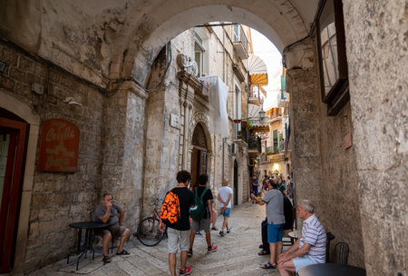 Bari, Puglia, Italy. August 2021. The charm of the alleys of the historic center, the old bari. People sitting on the side of the road chat under a stone arch. Tourists along the way.のeditorial素材