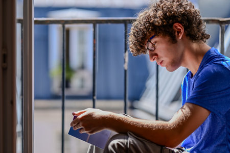 Nice shot of a young Caucasian man reading a book. He is sitting on an attic window, facing the sun....