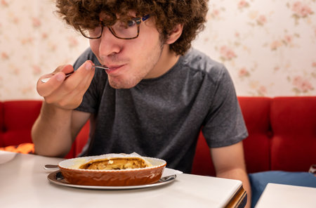 A cute close-up of a young Caucasian man eating a Viennese sweet specialty: apple strudel with custard. His eyes are amazed as he savors the bite.の写真素材