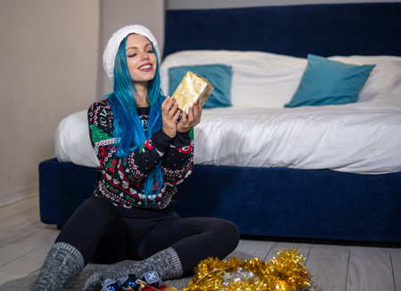 Portrait of a caucasian hipster woman in Christmas clothes. She is sitting next to the bed and smilingly holding a package. Expectation, surprise, happiness.の写真素材