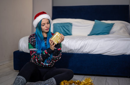 Portrait of a beautiful caucasian hipster woman in Christmas clothes. She is sitting next to the bed and smilingly holding a package. Expectation, surprise, happiness.の写真素材