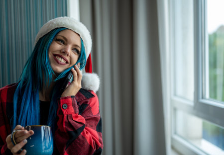 A hipster caucasian woman in Christmas clothes. She is talking on her cell phone while looking out the window, she has a cup of tea. Christmas,talking with friends, positivity and happiness.の写真素材