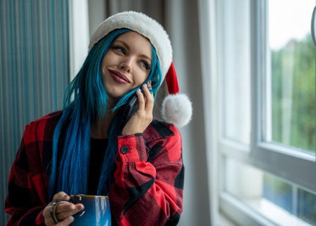 A beautiful hipster caucasian woman in Christmas clothes. She is talking on her cell phone while looking out the window, she has a cup of tea. Christmas,talking with friends, positivity and happiness.の写真素材