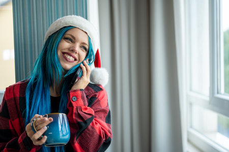 A beautiful hipster caucasian woman in Christmas clothes. She is talking on her cell phone while looking out the window, she has a cup of tea. Christmas,talking with friends, positivity and happiness.の写真素材
