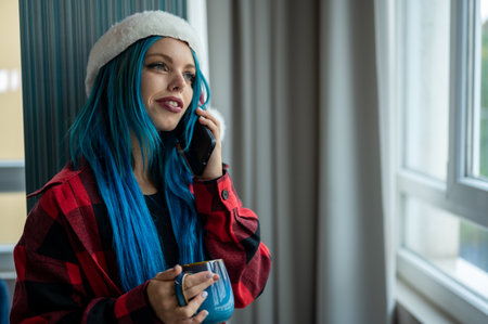 A hipster caucasian woman in Christmas clothes. She is talking on her cell phone while looking out the window, she has a cup of tea. Christmas,talking with friends, positivity and happiness.の写真素材