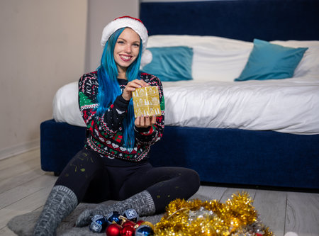 Portrait of a beautiful caucasian hipster woman in Christmas clothes. She is sitting next to the bed and smilingly holding a package. Expectation, surprise, happiness.の写真素材