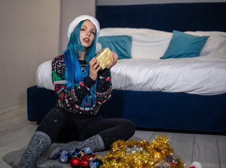 Portrait of a beautiful caucasian hipster woman in Christmas clothes. She is sitting next to the bed and smilingly holding a package. Expectation, surprise, happiness.の写真素材