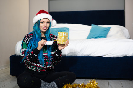 Portrait of a beautiful caucasian hipster woman in Christmas clothes. She is sitting next to the bed and smilingly unwrapping a package. Expectation, surprise, happiness.の写真素材