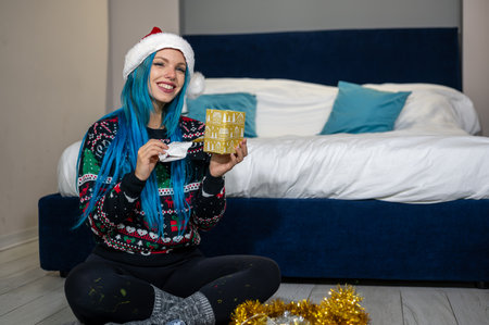 Portrait of a beautiful caucasian hipster woman in Christmas clothes. She is sitting next to the bed and smilingly unwrapping a package. Expectation, surprise, happiness.の写真素材