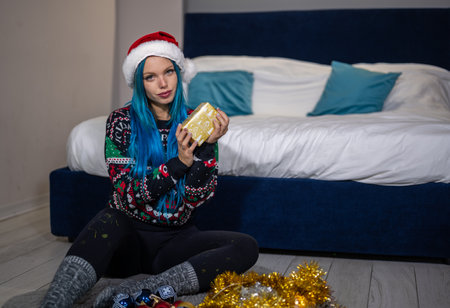 Portrait of a beautiful caucasian hipster woman in Christmas clothes. She is sitting next to the bed and smilingly holding a package. Expectation, surprise, happiness.の写真素材