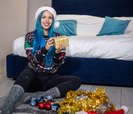Portrait of a beautiful caucasian hipster woman in Christmas clothes. She is sitting next to the bed and smilingly holding a package. Expectation, surprise, happiness.の写真素材