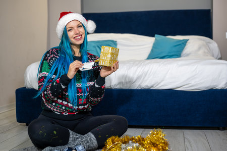 Portrait of a beautiful caucasian hipster woman in Christmas clothes. She is sitting next to the bed and smilingly unwrapping a package. Expectation, surprise, happiness.の写真素材