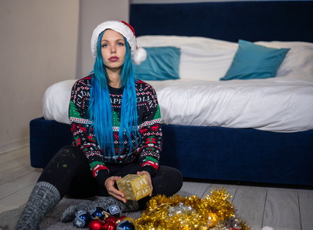 Portrait of a beautiful caucasian hipster woman in Christmas clothes. She is sitting next to the bed and smilingly holding a package. Expectation, surprise, happiness.の写真素材