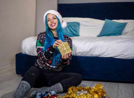 Portrait of a beautiful caucasian hipster woman in Christmas clothes. She is sitting next to the bed and smilingly holding a package. Expectation, surprise, happiness.の写真素材