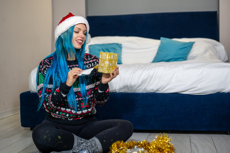 Portrait of a beautiful caucasian hipster woman in Christmas clothes. She is sitting next to the bed and smilingly unwrapping a package. Expectation, surprise, happiness.の写真素材