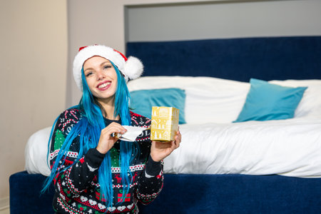 Portrait of a beautiful caucasian hipster woman in Christmas clothes. She is sitting next to the bed and smilingly unwrapping a package. Expectation, surprise, happiness.の写真素材
