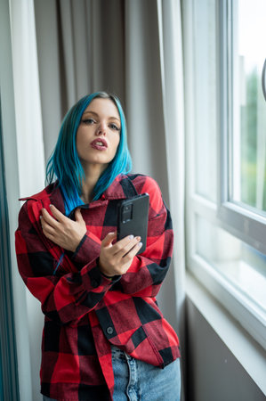 Vertical portrait of blue haired hipster woman standing in front of window taking selfie with her mobile phone. Looking at camera. Using technology.の写真素材