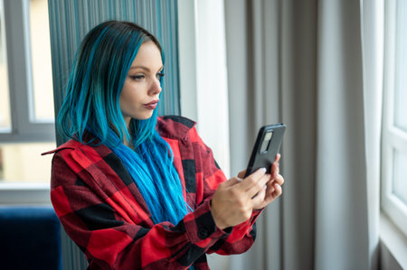 Portrait of beautiful blue haired caucasian hipster woman standing in front of window taking selfie with her mobile phone. Using technology.の写真素材