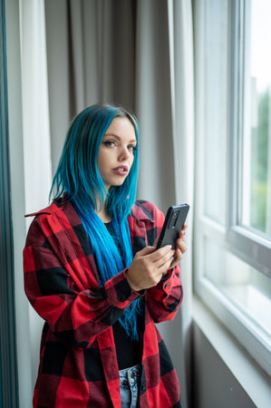 Vertical portrait of blue haired hipster caucasian woman standing in front of window taking selfie with her mobile phone. Looking at camera. Using technology.の写真素材